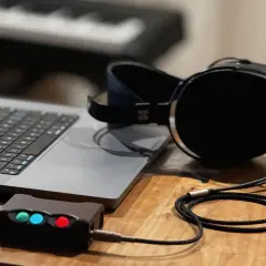Ein Laptop, ein Kopfhörerverstärker mit leuchtenden LEDs und ein Kopfhörer mit großem **headphone driver** liegen auf einem Holztisch.