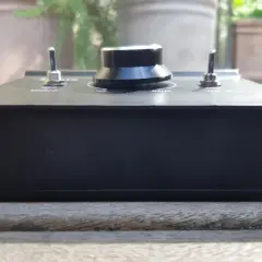 Schwarzes Audiogerät mit Drehknopf und Kippschaltern, das auf einer Holzoberfläche steht. Ein Hauch von grauem Rauschen in der Unschärfe des Hintergrunds.