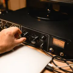 Hand dreht Regler an schwarzem Preamp mit VU-Meter, daneben Laptops und Kabel.