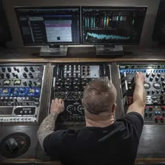 Mann mit Tattoos im Tonstudio, der an Reglern dreht, um einen perfekten pop mix zu kreieren. Monitore zeigen Wellenformen und Spektrumanalysen.