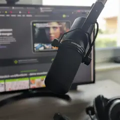 Mikrofon Shure SM7B vor einem Monitor mit Webseite, Kopfhörer daneben. Für klare Aufnahmen ohne noise floor.