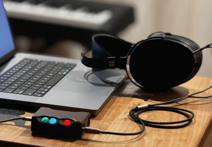Ein Laptop, ein Kopfhörerverstärker mit leuchtenden LEDs und ein Kopfhörer mit großem **headphone driver** liegen auf einem Holztisch.