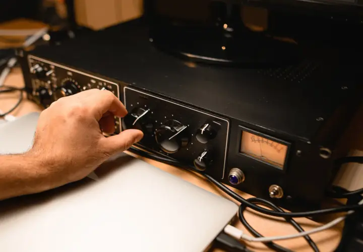Hand dreht Regler an schwarzem Preamp mit VU-Meter, daneben Laptops und Kabel.