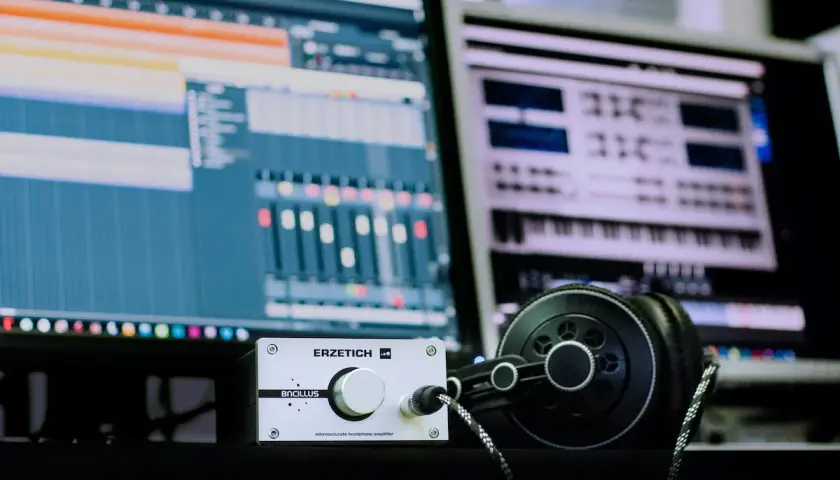 Kopfhörer und ein Verstärker liegen vor zwei Monitoren, die eine digitale Audio-Workstation zeigen. Hier wird Musik gemacht.