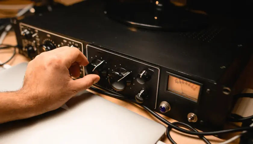 Hand dreht Regler an schwarzem Preamp mit VU-Meter, daneben Laptops und Kabel.