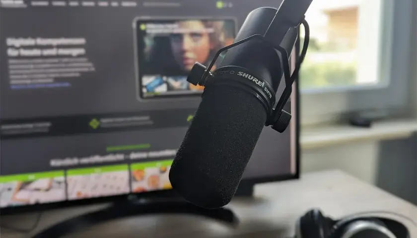 Mikrofon Shure SM7B vor einem Monitor mit Webseite, Kopfhörer daneben. Für klare Aufnahmen ohne noise floor.