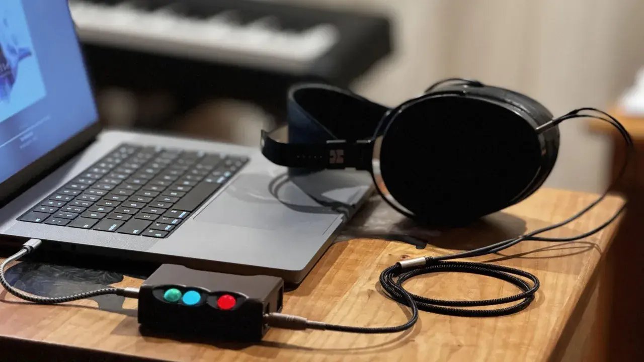 Ein Laptop, ein Kopfhörerverstärker mit leuchtenden LEDs und ein Kopfhörer mit großem **headphone driver** liegen auf einem Holztisch.