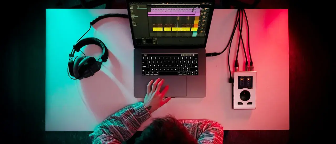 Person am Schreibtisch mit Kopfhörern, Laptop und Audio-Interface. Musikproduktion in 24 bit 48 khz.