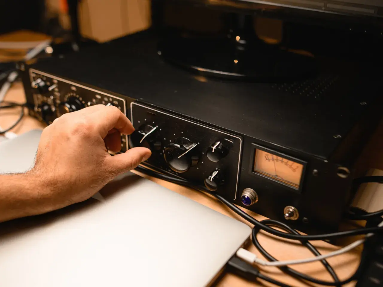Hand dreht Regler an schwarzem Preamp mit VU-Meter, daneben Laptops und Kabel.