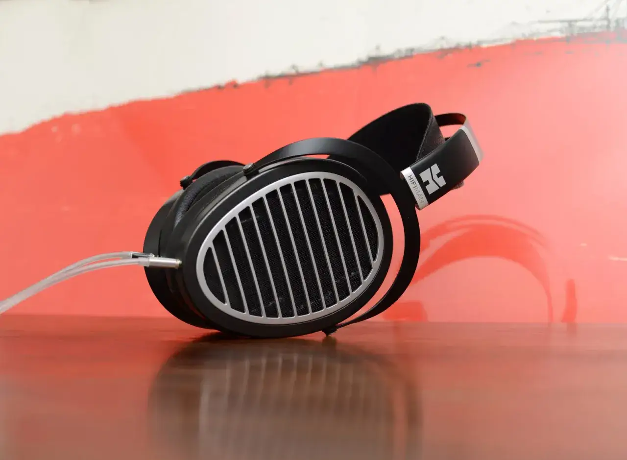 Hifiman Ananda im Test: Hochwertige Kopfhörer mit offenem Design auf einem Holztisch vor rotem Hintergrund.