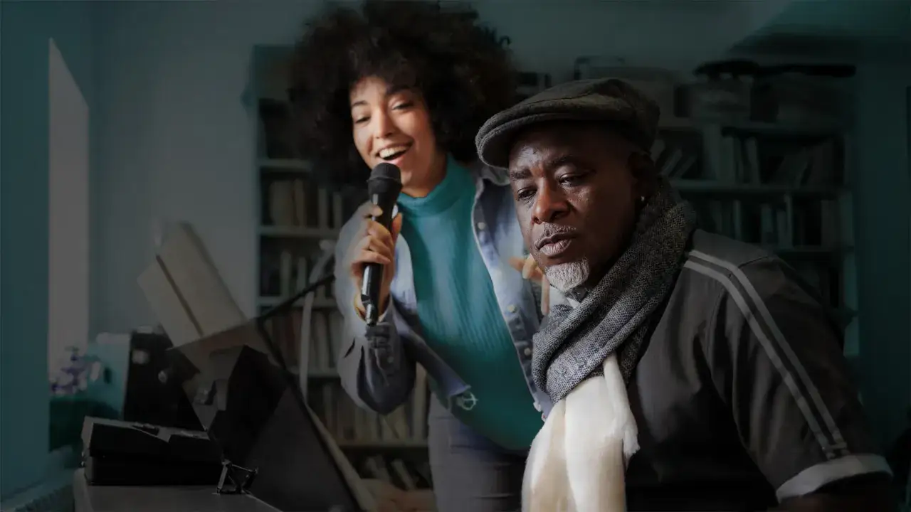 Junge Frau singt ins Mikrofon, während ein älterer Mann mit Mütze und Schal zuhört. Ein Klavier steht im Vordergrund. Der Moment des formant shifting, wenn Musik verbindet.