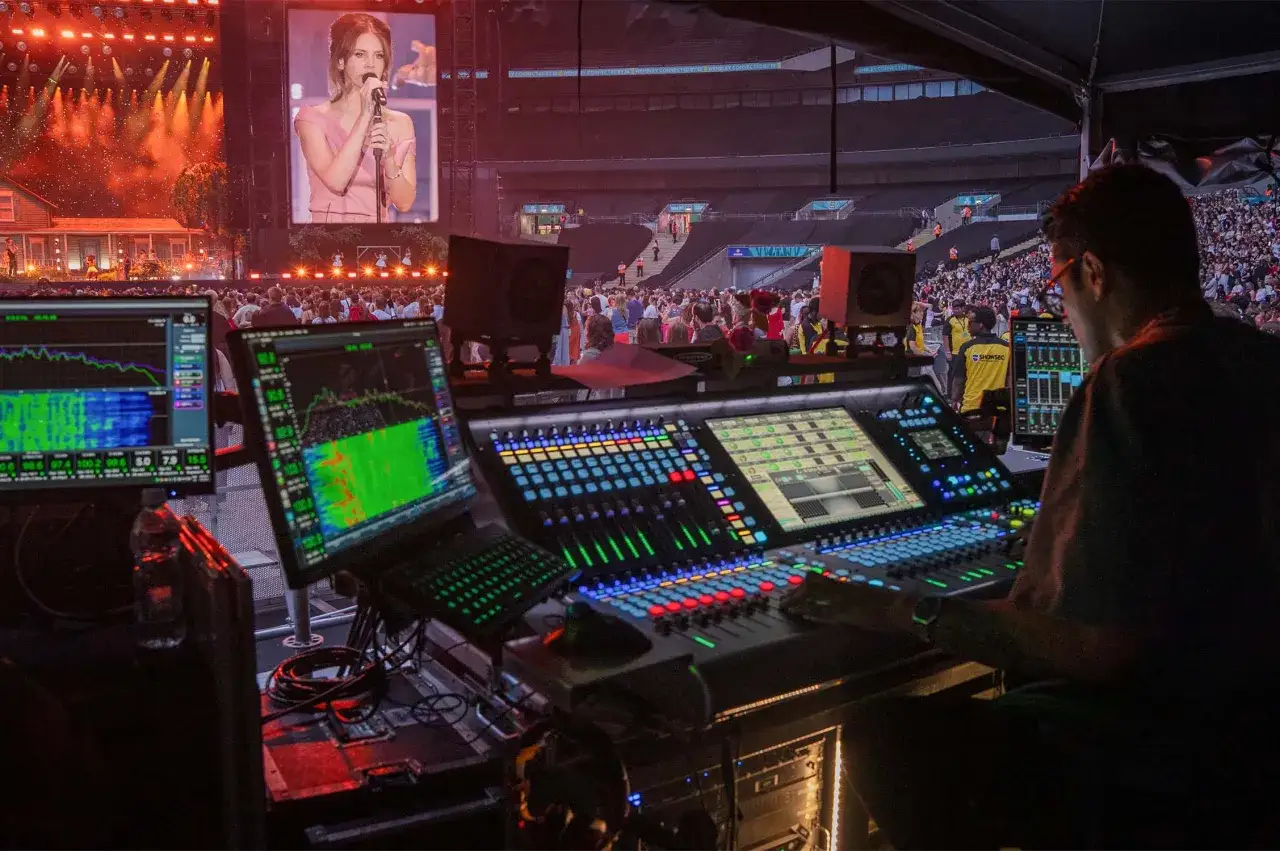 Tontechniker mischt live sound bei einem Konzert. Auf den Bildschirmen sind Frequenzanalysen zu sehen, w&auml;hrend im Hintergrund die K&uuml;nstlerin auf einer riesigen Leinwand performt.